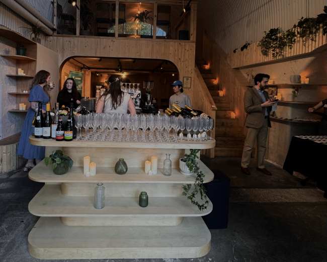 The image shows a modern bar interior with an organized display of wine bottles and champagne flutes, while patrons socialize and staff serve drinks in a spacious, well-lit space.