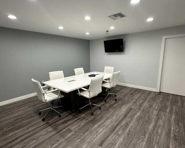 A modern, minimalist conference room features a rectangular table surrounded by white chairs, with a television mounted on the wall and recessed lighting above.
