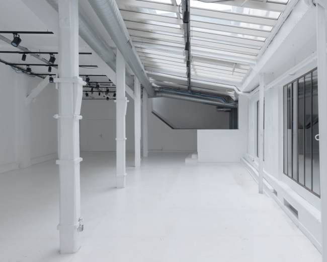 The image shows a spacious, bright room with white walls and a glossy floor, featuring large windows and a minimalist design.