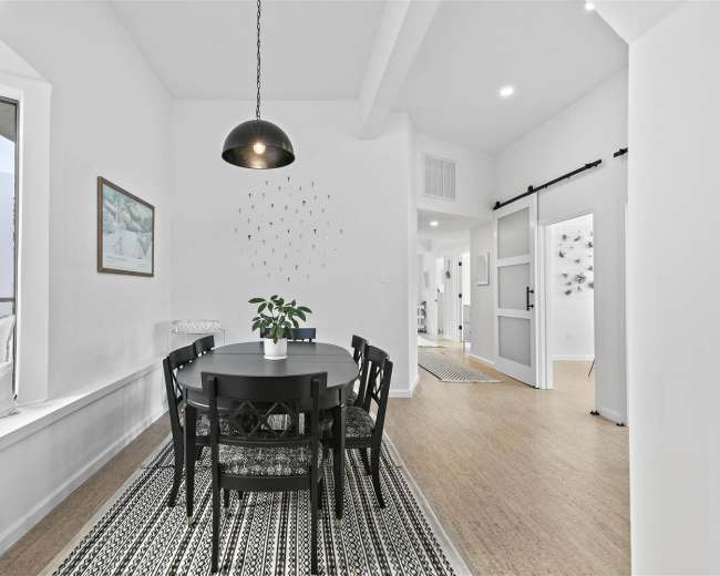 A dining area features a round black table surrounded by six chairs, with large windows and a potted plant on the table.