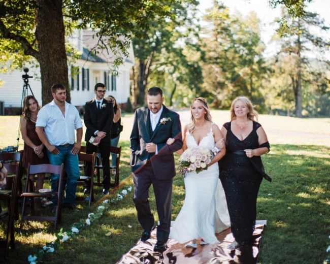 A bride is walking down an aisle flanked by her mother and a man, while guests are seated on either side.
