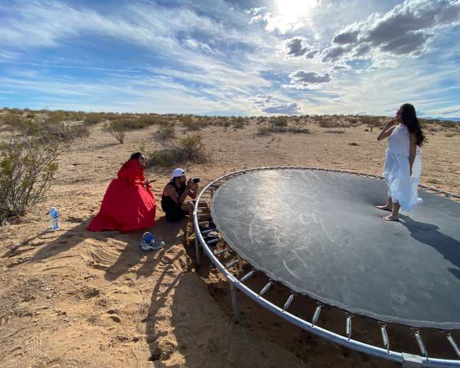 A model poses on a trampoline in a desert setting while two photographers, one in a red dress and the other in black, capture the scene.