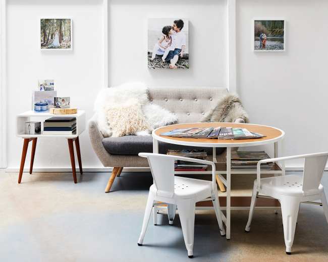 A modern living room featuring a gray sofa, a round wooden coffee table with magazines, and two white chairs, all against a backdrop of white walls adorned with framed photos.