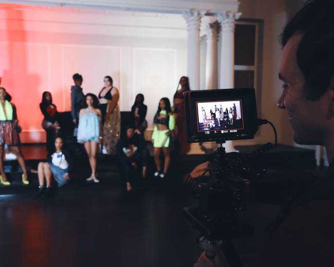 A photographer captures a group of models posing on a stage while using a camera mounted on a stabilizer.