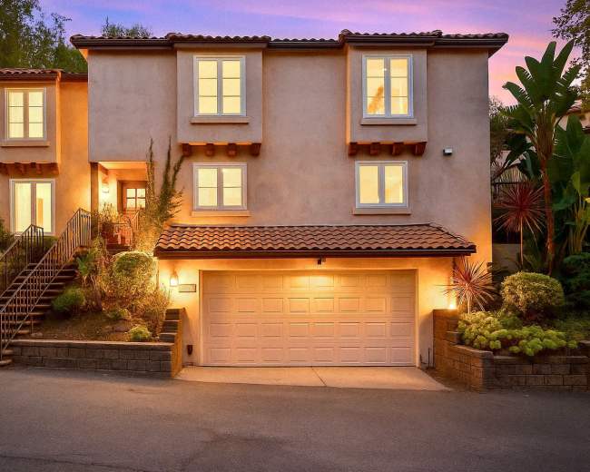 The image shows a two-story house with a tile roof, featuring a garage and a landscaped front yard, illuminated at dusk.