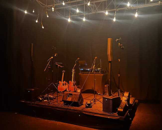 A dimly lit stage set up with guitars, microphones, and sound equipment surrounded by hanging lights.