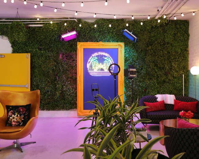 The image shows a stylish indoor space featuring a lush green wall, neon signage, modern furniture, and hanging lights.
