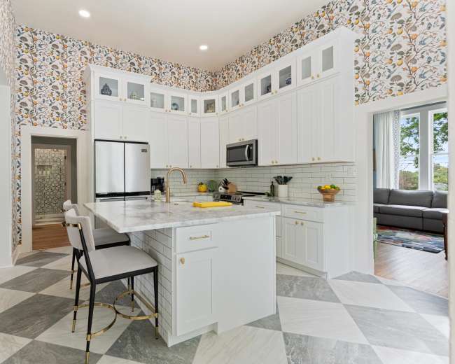 The image shows a modern kitchen with white cabinetry, a marble island, and floral wallpaper, featuring stainless steel appliances and a checkered floor.