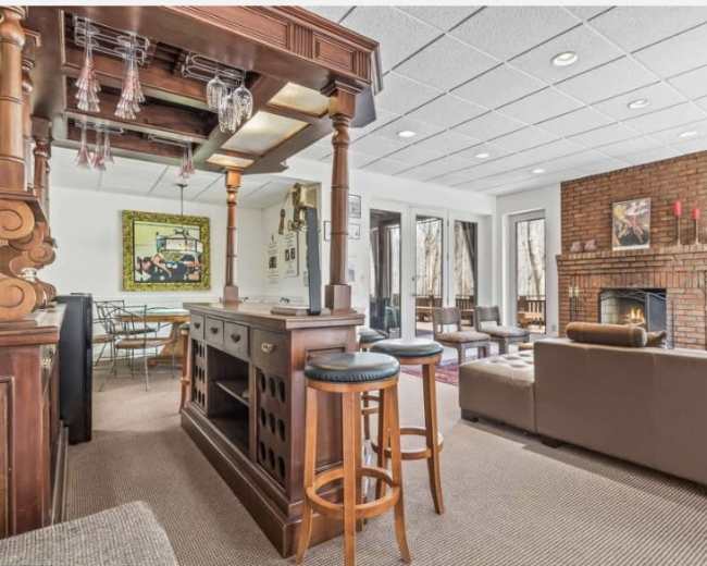 The image depicts a living space with a wooden bar area, seating, a fireplace, and large windows allowing natural light.