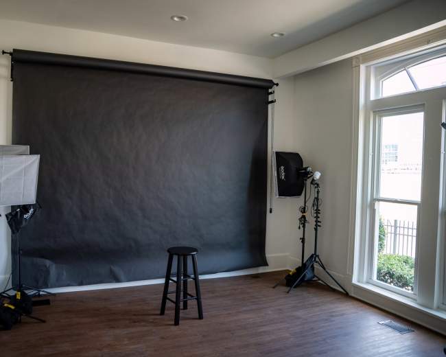 The image shows a photography studio with a dark backdrop, a black stool at the center, and lighting equipment positioned around the room.