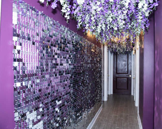 A hallway is adorned with a purple wall and a mirrored mosaic on one side, while artificial flowers hang from the ceiling.