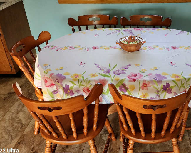 A round wooden dining table with a floral tablecloth and a bowl in the center is surrounded by six wooden chairs.