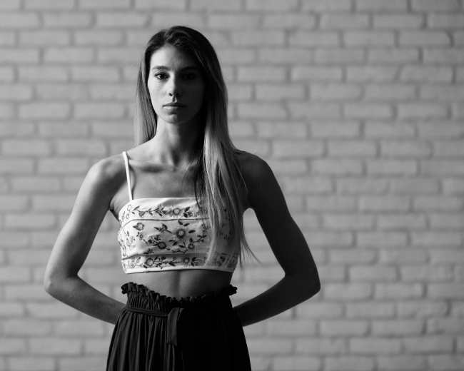 A woman stands with her hands behind her back in front of a textured brick wall.