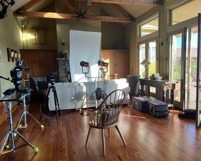 A living room setup for a video shoot, featuring cameras on tripods, a large white reflector, and a wooden chair positioned in the center.