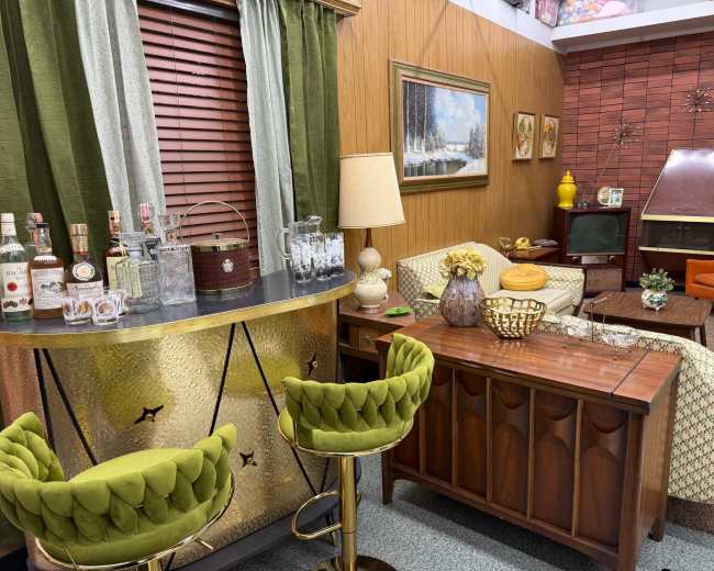 The image shows a retro living room and bar area featuring vintage furniture, a television, and decorative items typical of mid-20th century design.