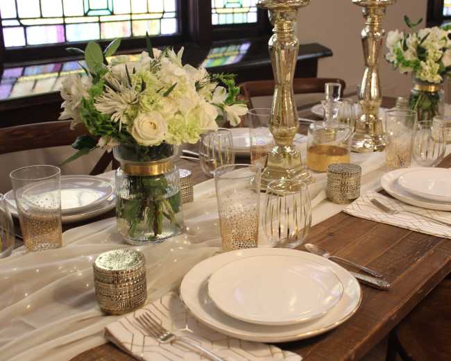 A dining table is set with white plates, floral centerpieces, glassware, and lit candles, all arranged on a white table runner.