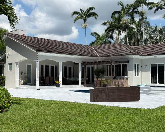 The image shows a modern house with a spacious patio area, palm trees in the background, and a small pool adjacent to the outdoor seating.