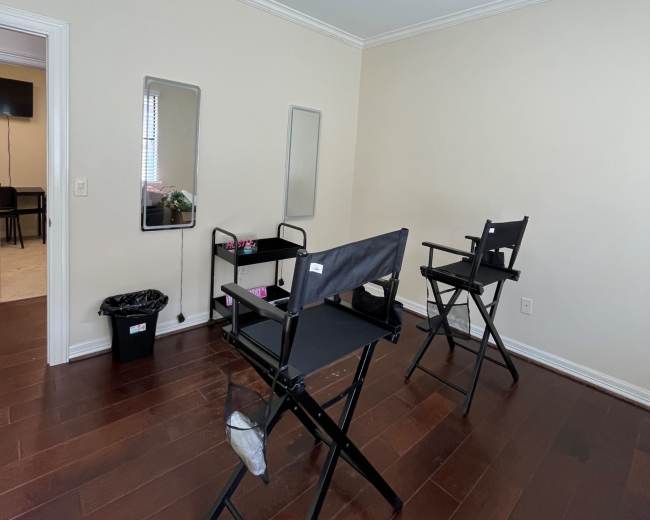 The image shows a small, well-lit room with two black director's chairs, a table with items on it, and a waste bin, all positioned on wooden flooring.