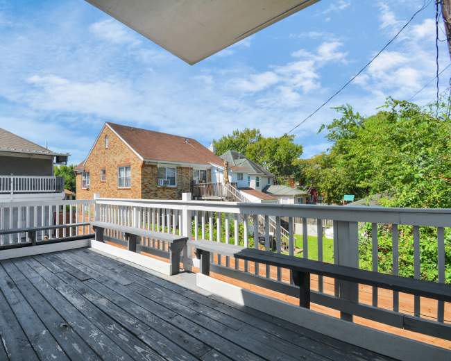 The image shows a wooden deck with benches, overlooking nearby houses and a green area under a partly cloudy sky.