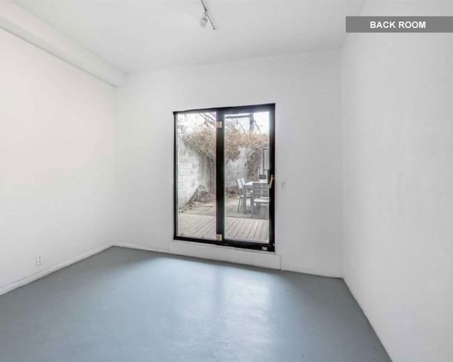 The image shows a minimally furnished room with white walls and a large window overlooking a deck area.