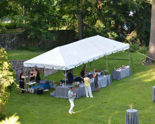A tented outdoor event features several tables with drinks and food, surrounded by trees and a grassy area.