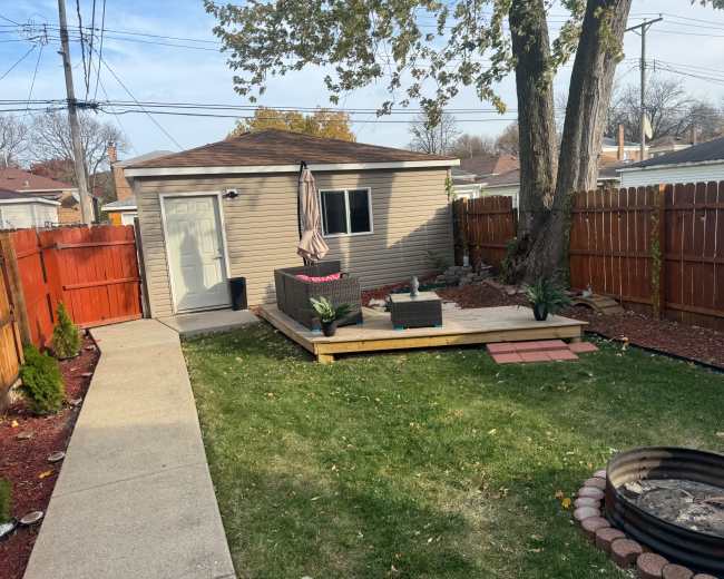 The image shows a backyard with a small shed, a patio area with outdoor furniture, and a fire pit surrounded by landscaping.