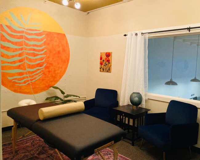 A treatment room featuring two massage tables, a blue armchair, a small side table, and a large orange wall mural of a leaf.