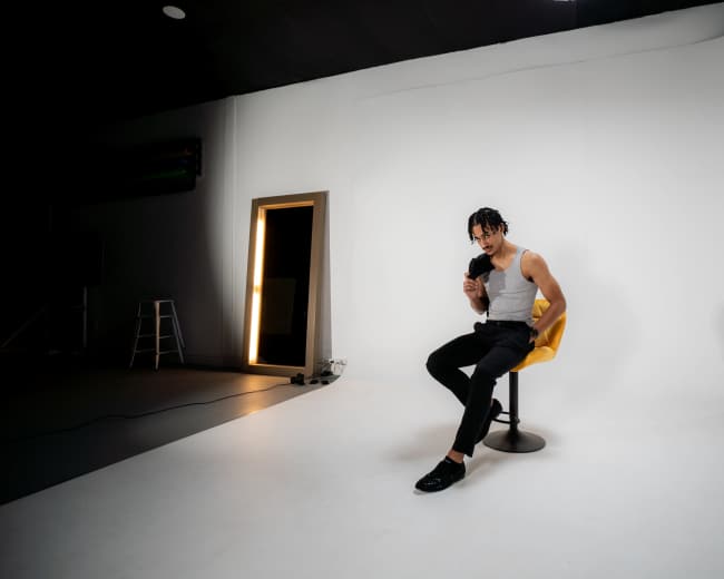A person with braided hair sits on a yellow chair in a studio with a white backdrop.