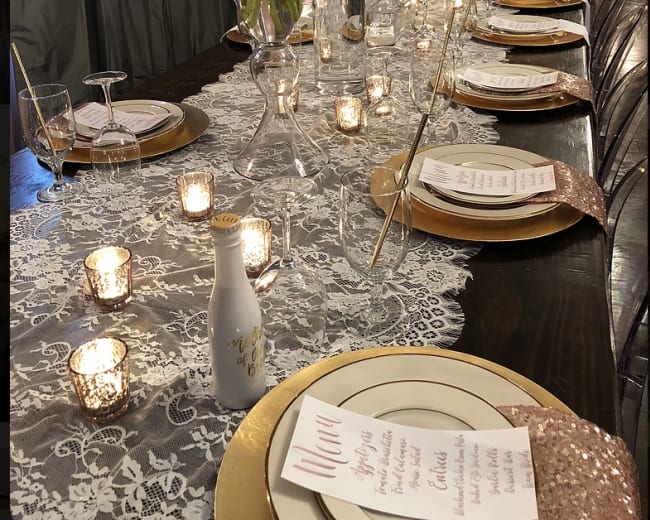 A long dining table is elegantly set with gold plates, white floral centerpieces, and lit candles alongside printed menus.