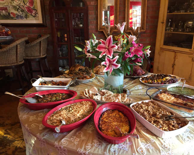 A variety of dishes, including casseroles and breads, are arranged on a table decorated with a vase of lilies and a tablecloth.
