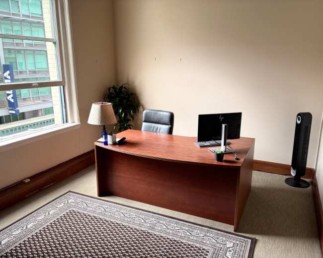 The image shows a simple office space with a wooden desk, a black office chair, a computer monitor, a small lamp, a plant, and a space heater, all against a plain wall and a window with city views.