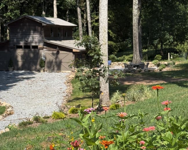 A gravel driveway leads to a wooden garage surrounded by trees, with a garden of colorful flowers in the foreground.