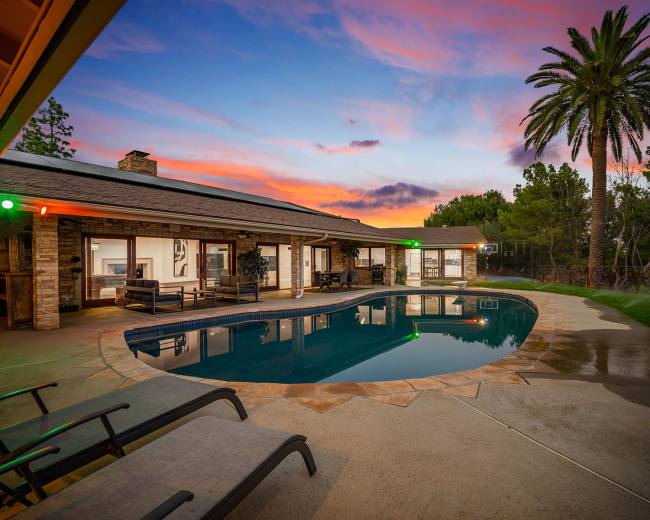 A modern home with a swimming pool reflects a vibrant sunset, surrounded by palm trees and outdoor seating.