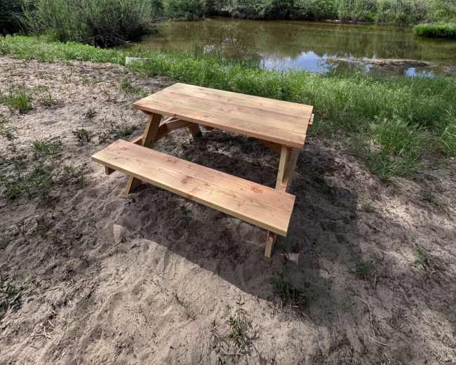 A wooden picnic table and bench sit on sandy ground near a calm pond surrounded by green vegetation.