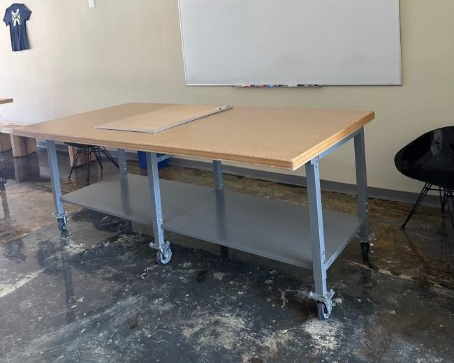 The image shows a large worktable on wheels in a room with a whiteboard and a few chairs.
