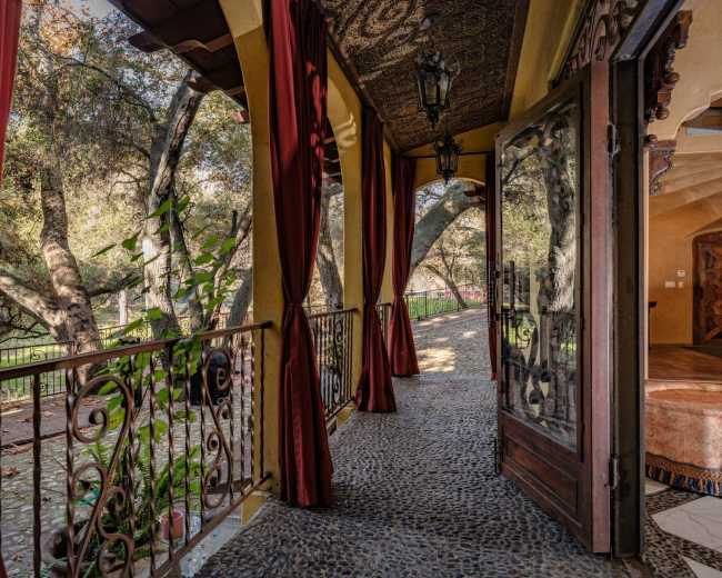 The image shows a balcony with a stone floor and decorative iron railings, framed by flowing red curtains and offering a view of a lush outdoor area.