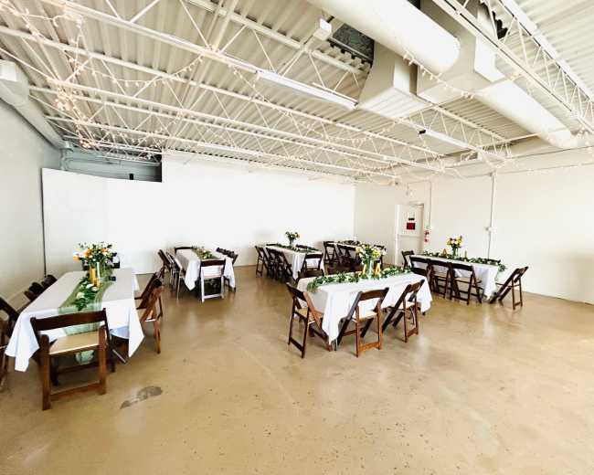 A spacious event room features several tables with white tablecloths and flower centerpieces arranged around a pale concrete floor.