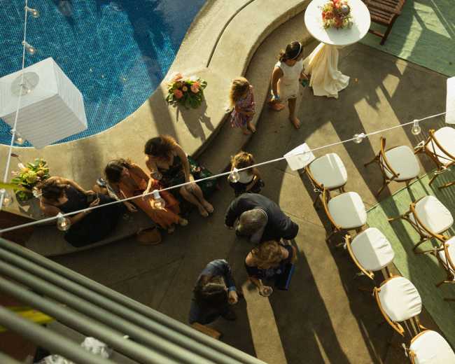 A group of people socializes around a poolside area adorned with flowers and string lights.