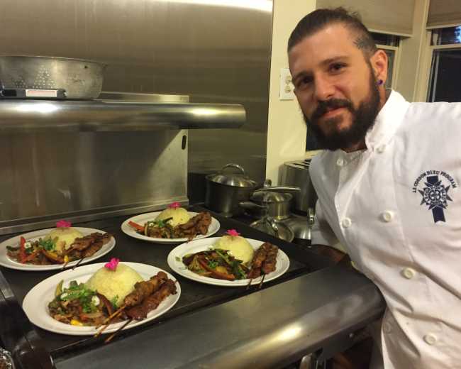 A chef stands beside four plated dishes featuring skewered meats, rice, and vegetables, each garnished with a flower.