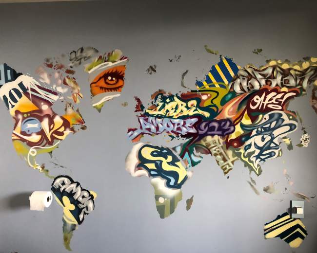 The image shows a wall mural of a world map covered in colorful graffiti and paint.