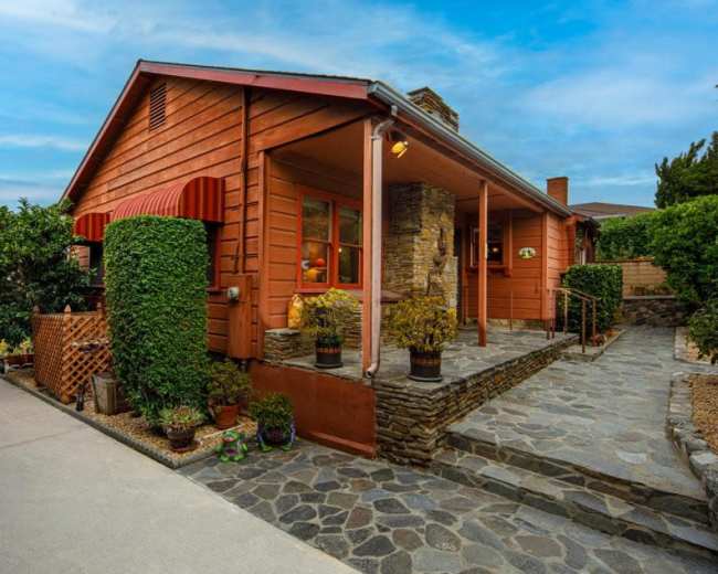A wooden house with a stone pathway and well-maintained shrubs is set against a clear blue sky.