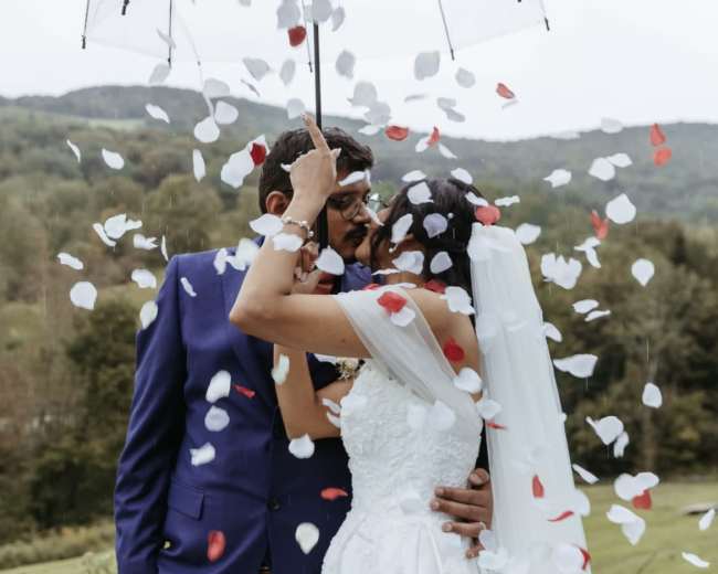 A couple stands under a clear umbrella while petals fall around them in a lush outdoor setting.