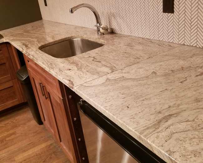 The image shows a kitchen corner featuring wooden cabinetry, a stainless steel sink, and a granite countertop with a herringbone-patterned backsplash.