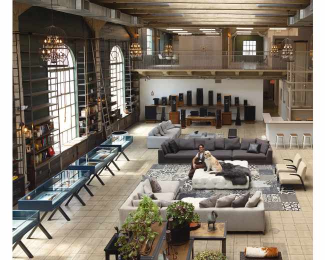 The image shows a spacious industrial-style loft with high ceilings, large windows, multiple seating areas, and various plants arranged on long tables.