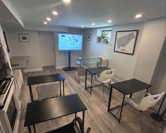 The image shows a small classroom-like setting with several black tables and chairs arranged around a large screen displaying a map.