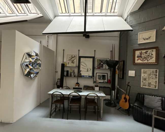 The image shows a spacious studio with a large skylight, featuring a table with chairs, artistic wall decor, and a guitar in the corner.