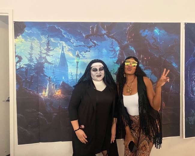 Two women pose for a photo in front of a large, dark-themed mural depicting a spooky landscape with a castle and trees.