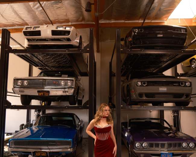 A woman in a red dress stands in a garage filled with classic cars displayed on multiple levels.