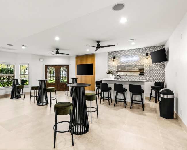 The image shows a modern indoor lounge area featuring high-top tables, a bar with stools, and large windows allowing natural light.