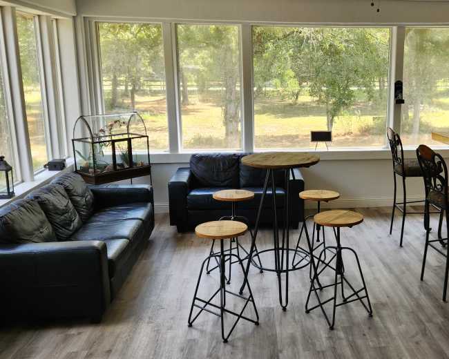 The image shows a sunlit lounge area with a leather couch, a round wooden table surrounded by four high stools, and a bar with a few bottles visible in the background.
